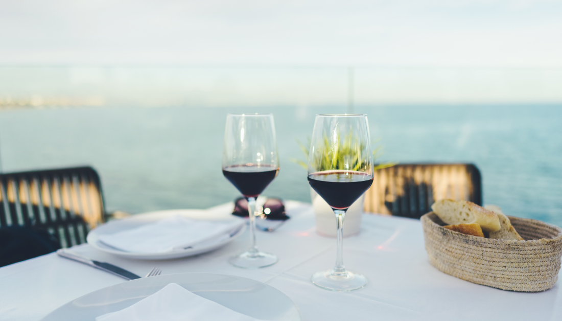 Two glass of red wine sit on a white table with an ocean setting behind